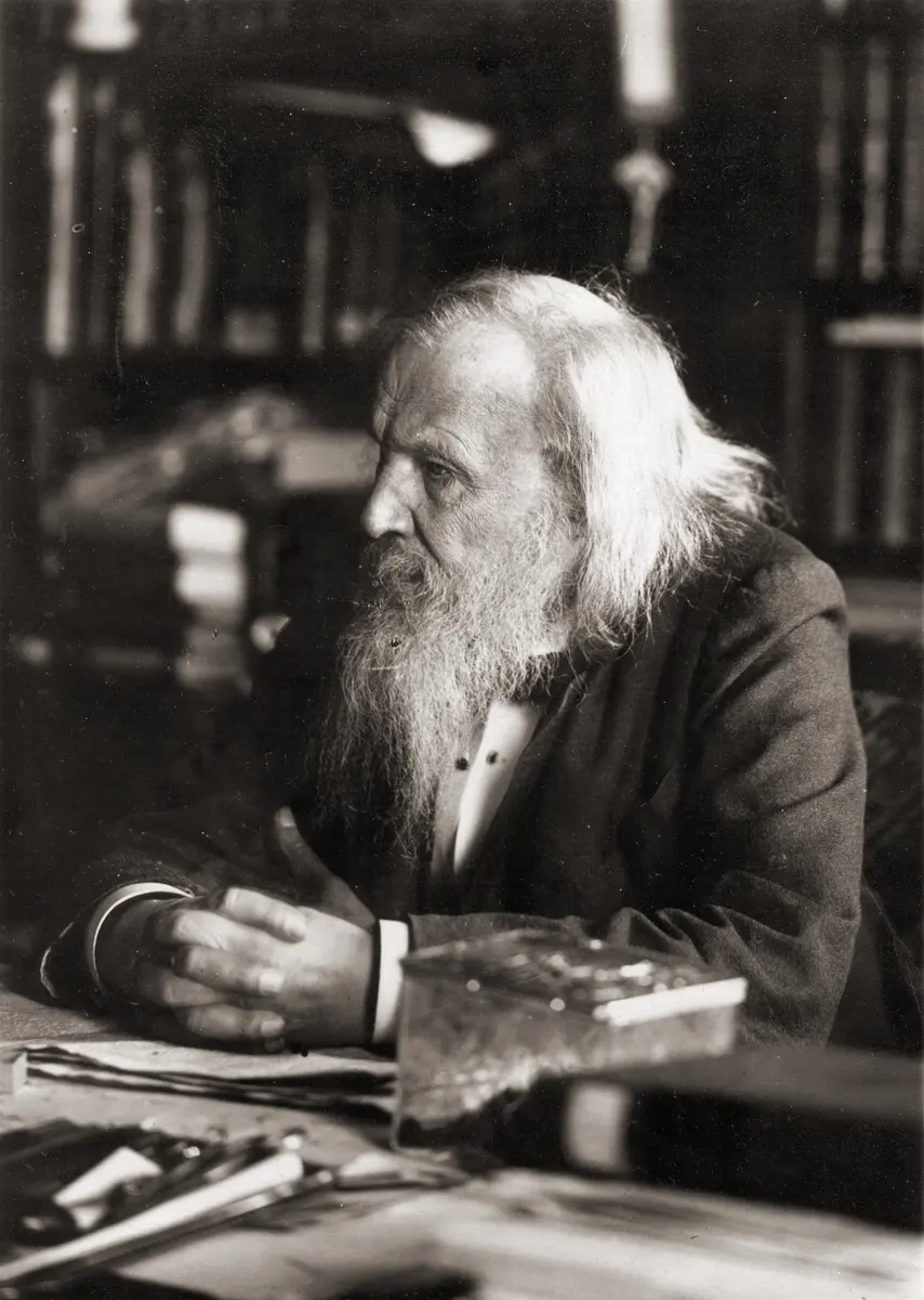 An old photo of an elderly man with long white hair and a long white beard, sitting at a desk with papers, in front of shelves of books