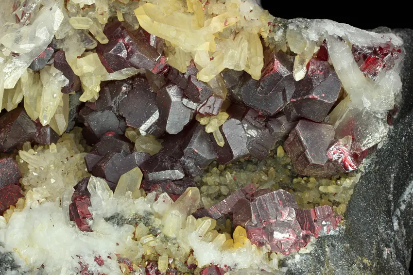 A close-up of a rock covered in blocky, dark red crystals with bright red edges, among pale yellow and white pointed quartz crystals