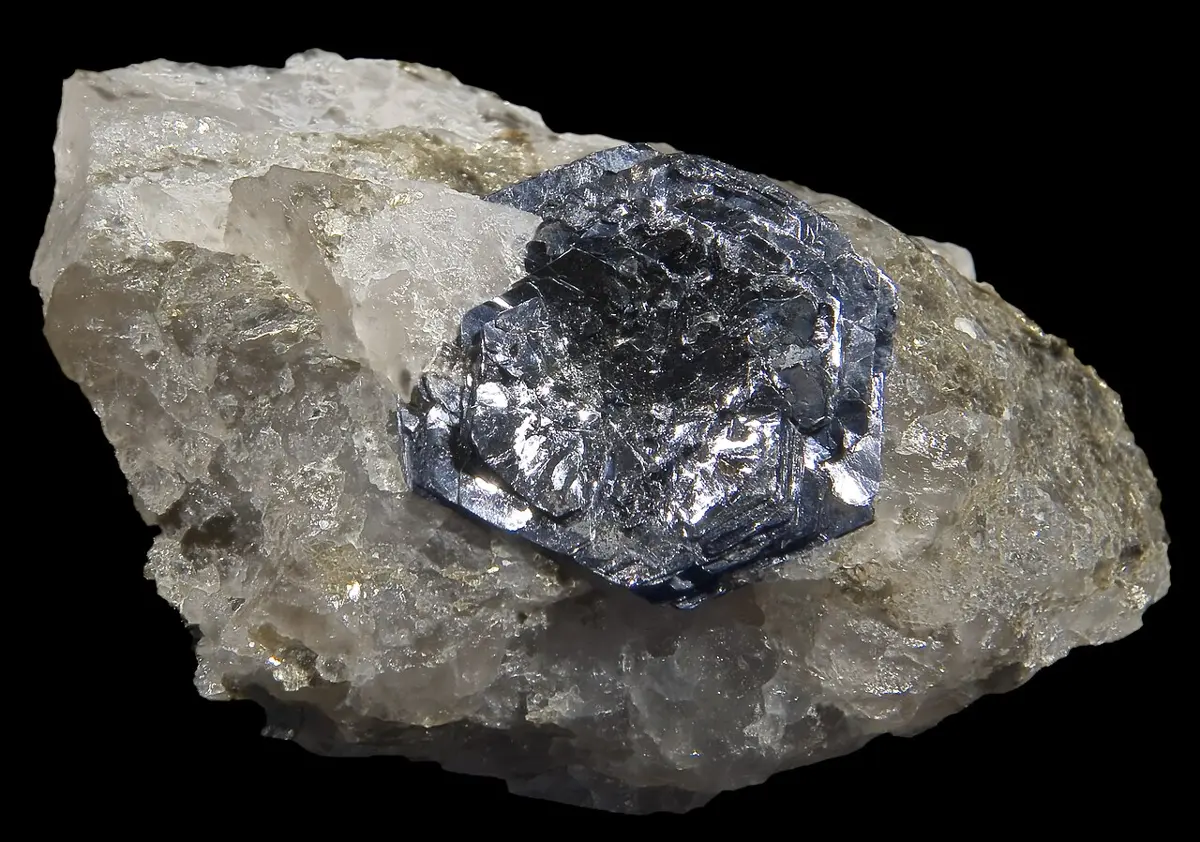 A flat, six-sided, shiny bluish-gray crystal sitting on a lump of pale gray quartz