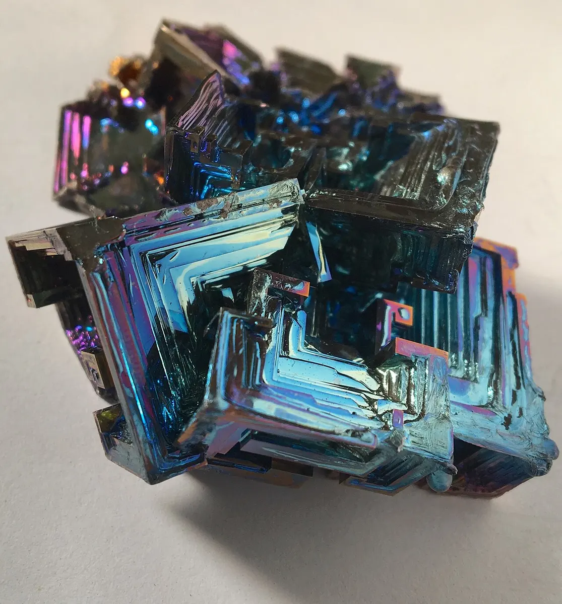 A cluster of bismuth crystals shaped like stepped, hollow squares, shining blue, purple and pink, on a white surface
