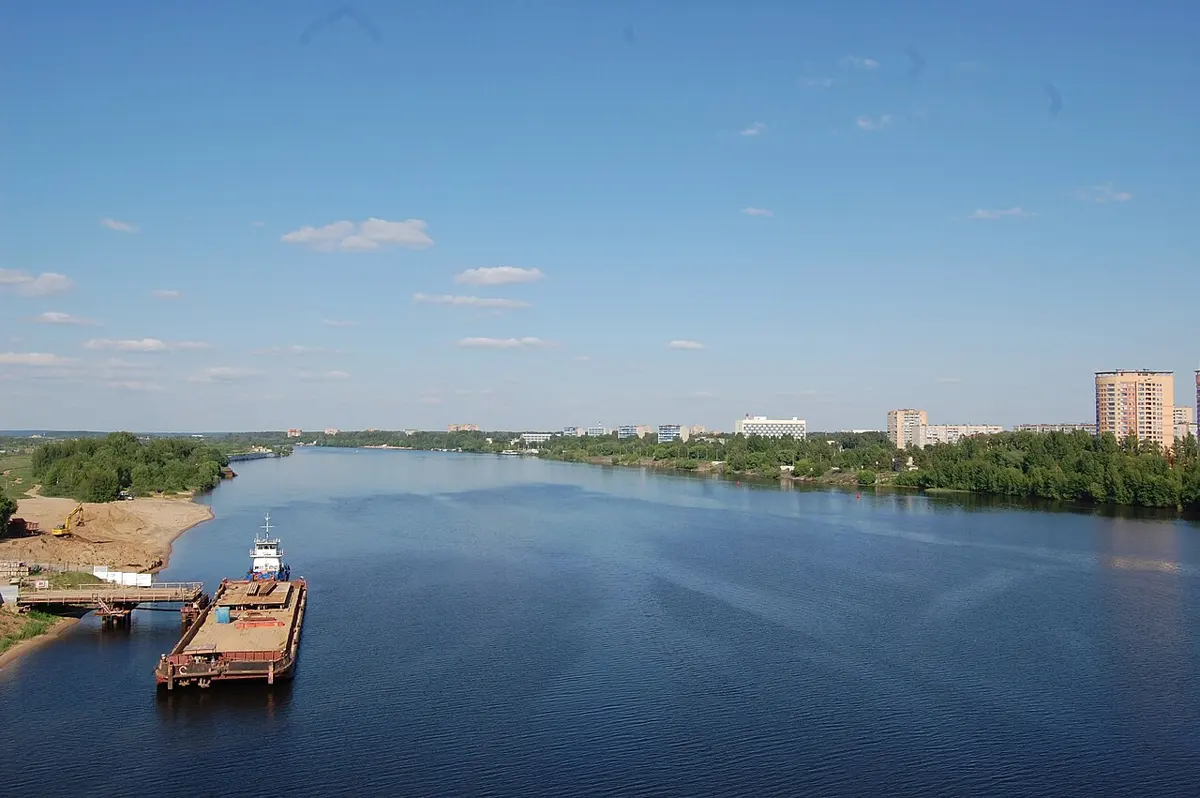 A wide blue river under a clear sky, seen from high up on a bridge; a barge is moored at a jetty on the left bank, and apartment blocks rise beyond the trees on the right