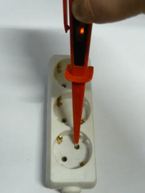 A red screwdriver tester pushed into one hole of a white power strip, a finger on its end, and a small orange light glowing in its handle