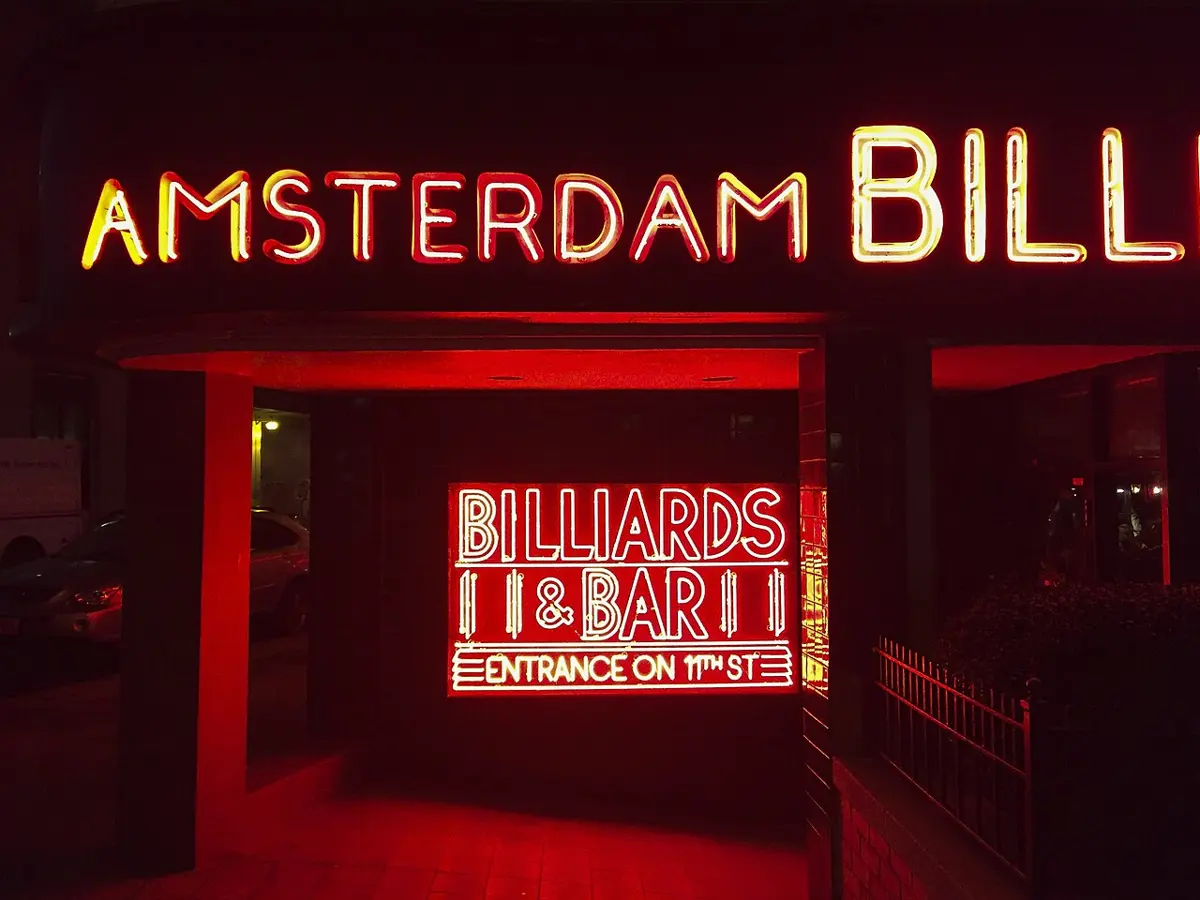 The entrance of a building at night, with the words Amsterdam Billiards along the top and Billiards and Bar in a window, all in glowing red tubes