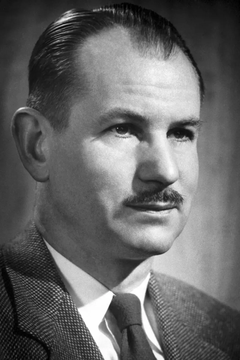 A black-and-white portrait of a man with slicked-back hair and a small mustache, in a tweed jacket and tie