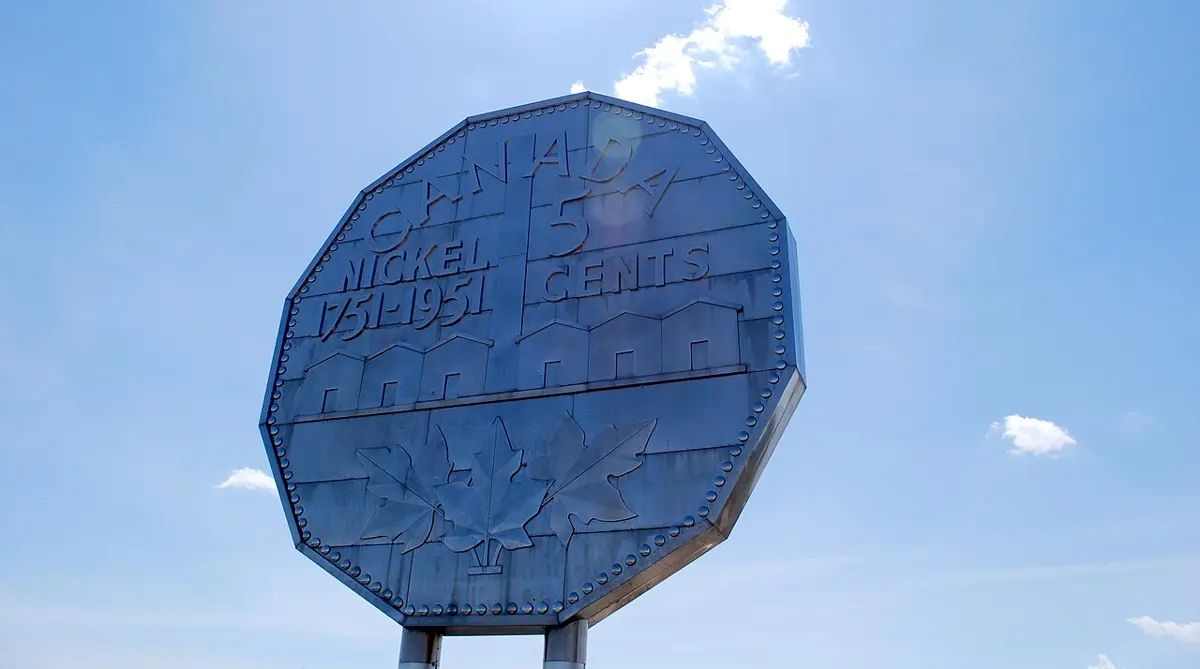A huge twelve-sided metal coin on posts against a blue sky, stamped Canada, Nickel 1751–1951 and 5 cents, with a row of buildings and maple leaves