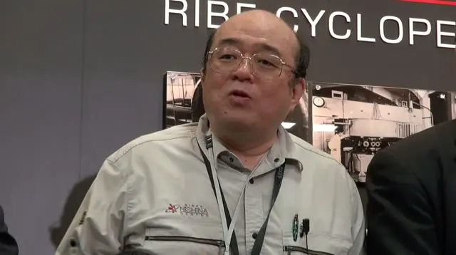 A man with glasses and short gray hair, in a light gray work jacket with a lanyard round his neck, speaking in front of a large photo of accelerator machinery