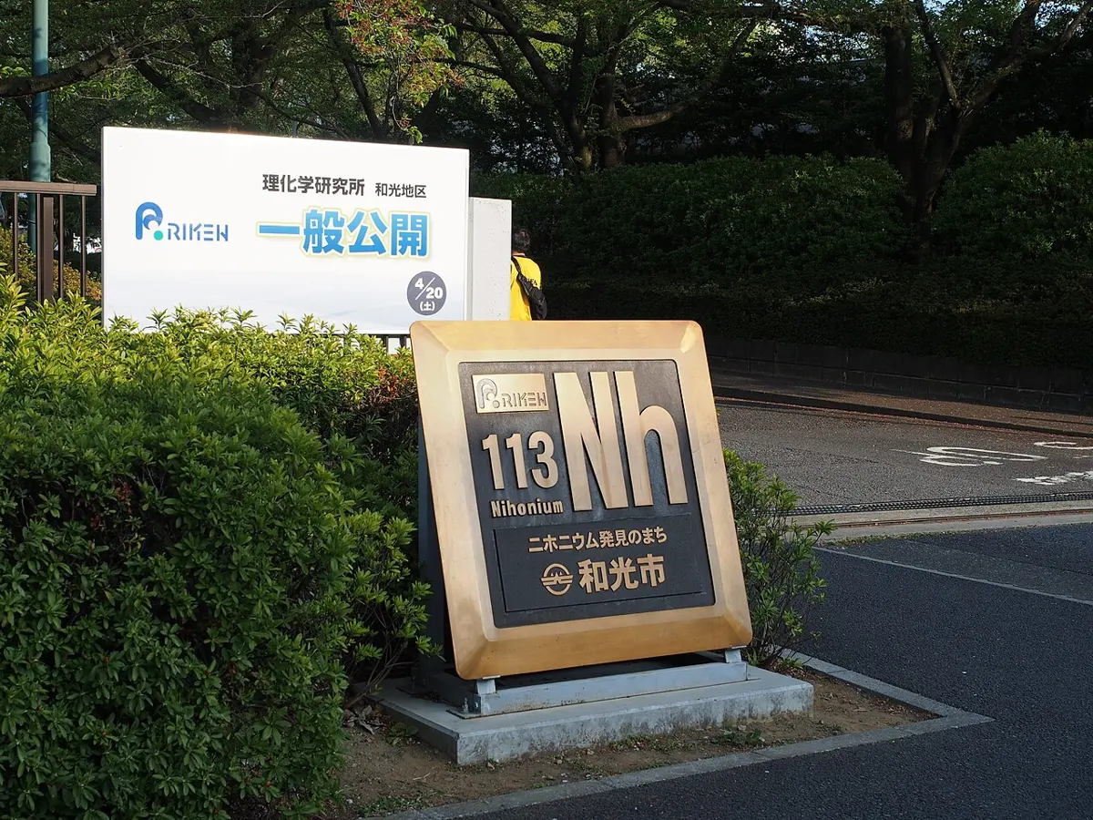 A bronze-colored plaque on a stand beside a hedge, showing a large “Nh” with “113” above it, in front of a sign for RIKEN and a road leading in