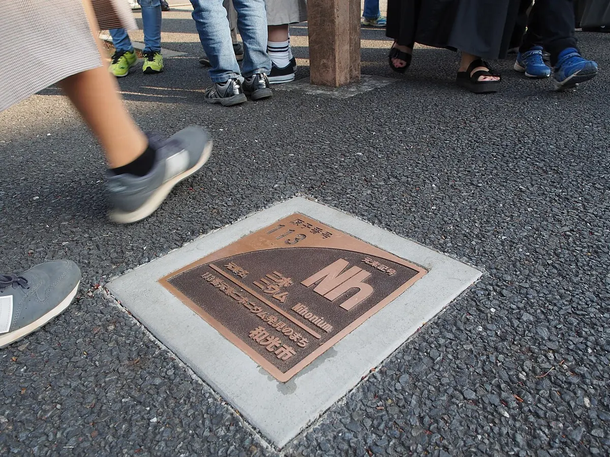 A bronze-colored plate set into a gray paved street, showing the symbol Nh with its number and name, with people’s feet walking past