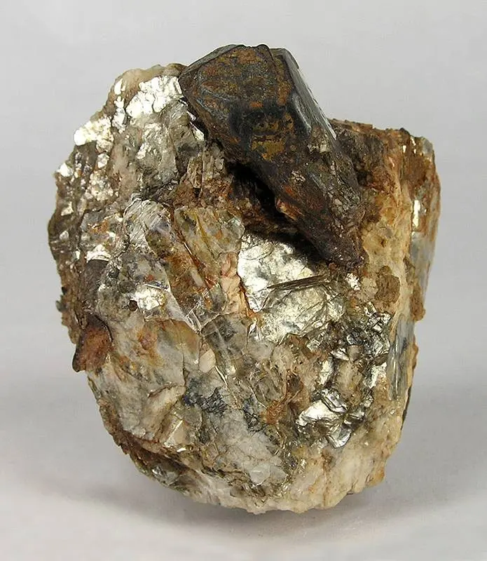 A dark brown crystal with flat faces sitting on top of a lump of pale rock full of shiny, silvery flakes