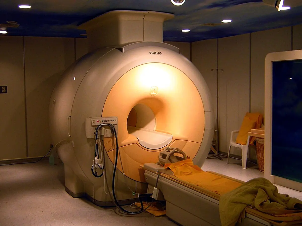 A large, rounded, cream-colored machine with a round tunnel through its middle, lit from inside, and a patient bed with yellow cushions in front of it