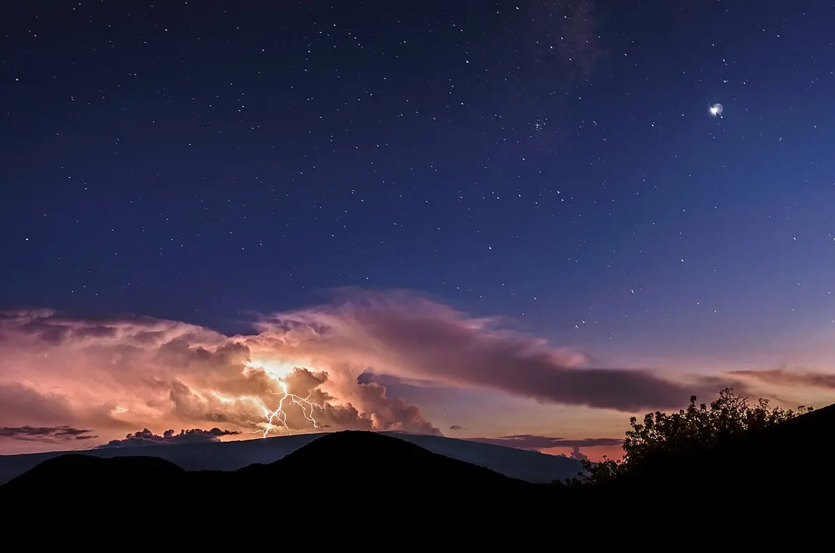 A starry evening sky over dark hills; far away, a branching bolt of lightning lights up a bank of pink clouds