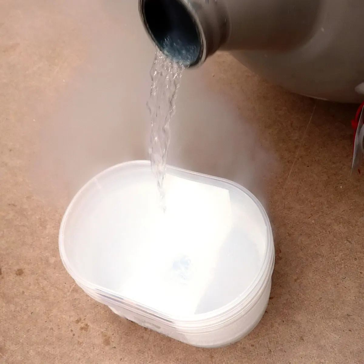 A clear liquid pouring from the neck of a metal flask into a white plastic tub, with white mist spilling around it
