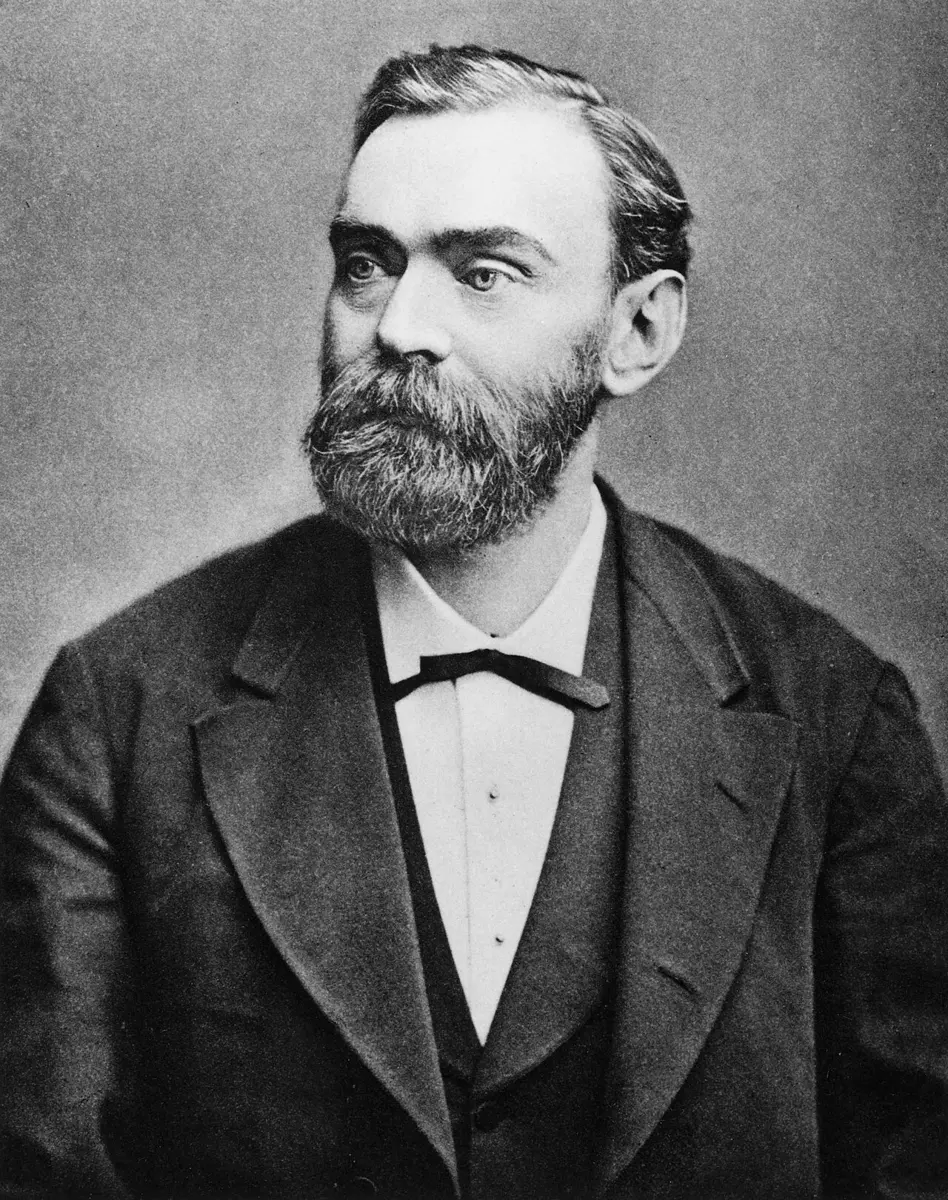 A black-and-white portrait of a man with dark, combed-back hair and a full beard, in a dark suit and bow tie
