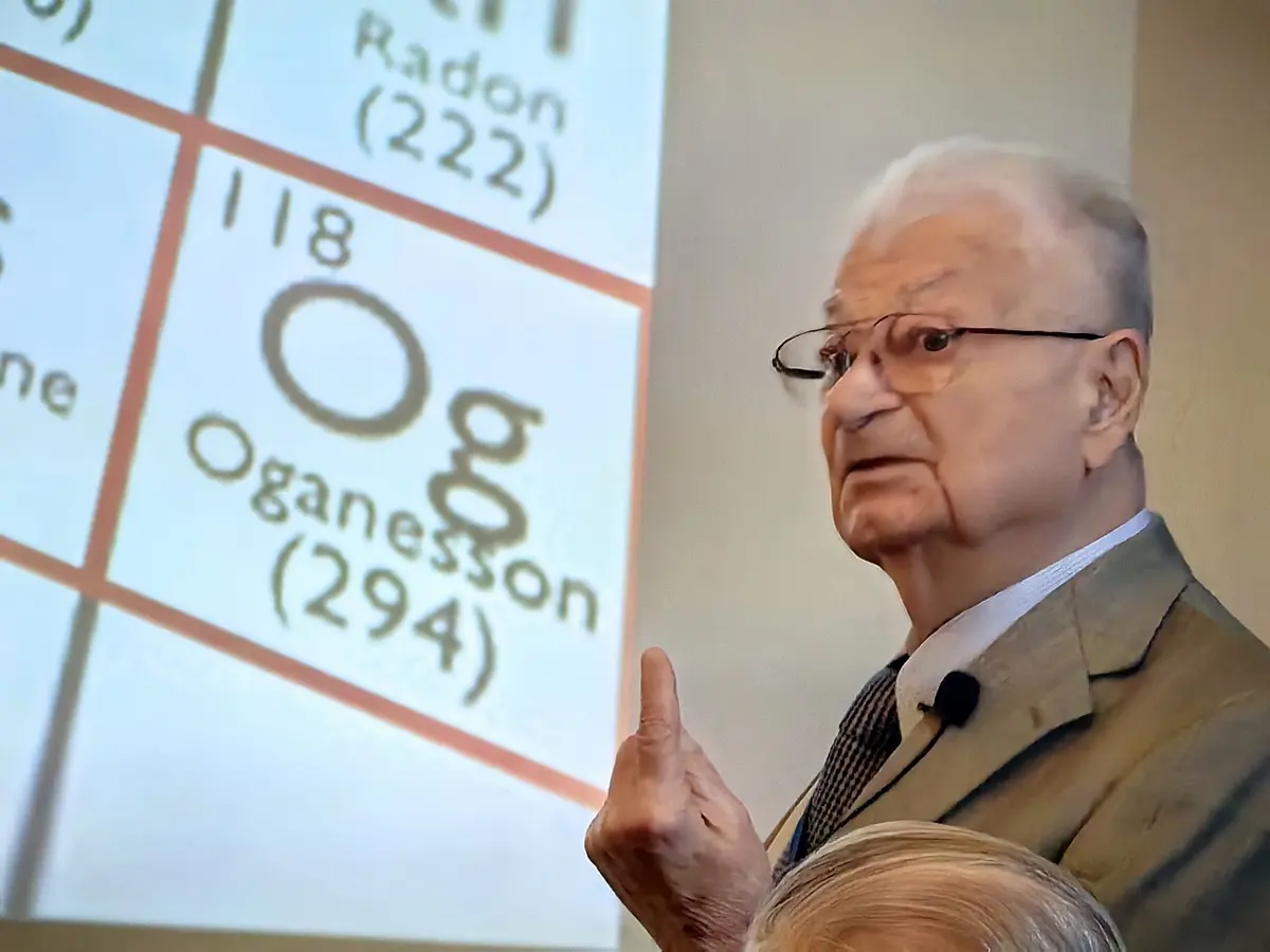 An elderly man with white hair and glasses, in a jacket and tie, speaks with one finger raised, in front of a screen showing the box for element 118, Og, oganesson, below radon