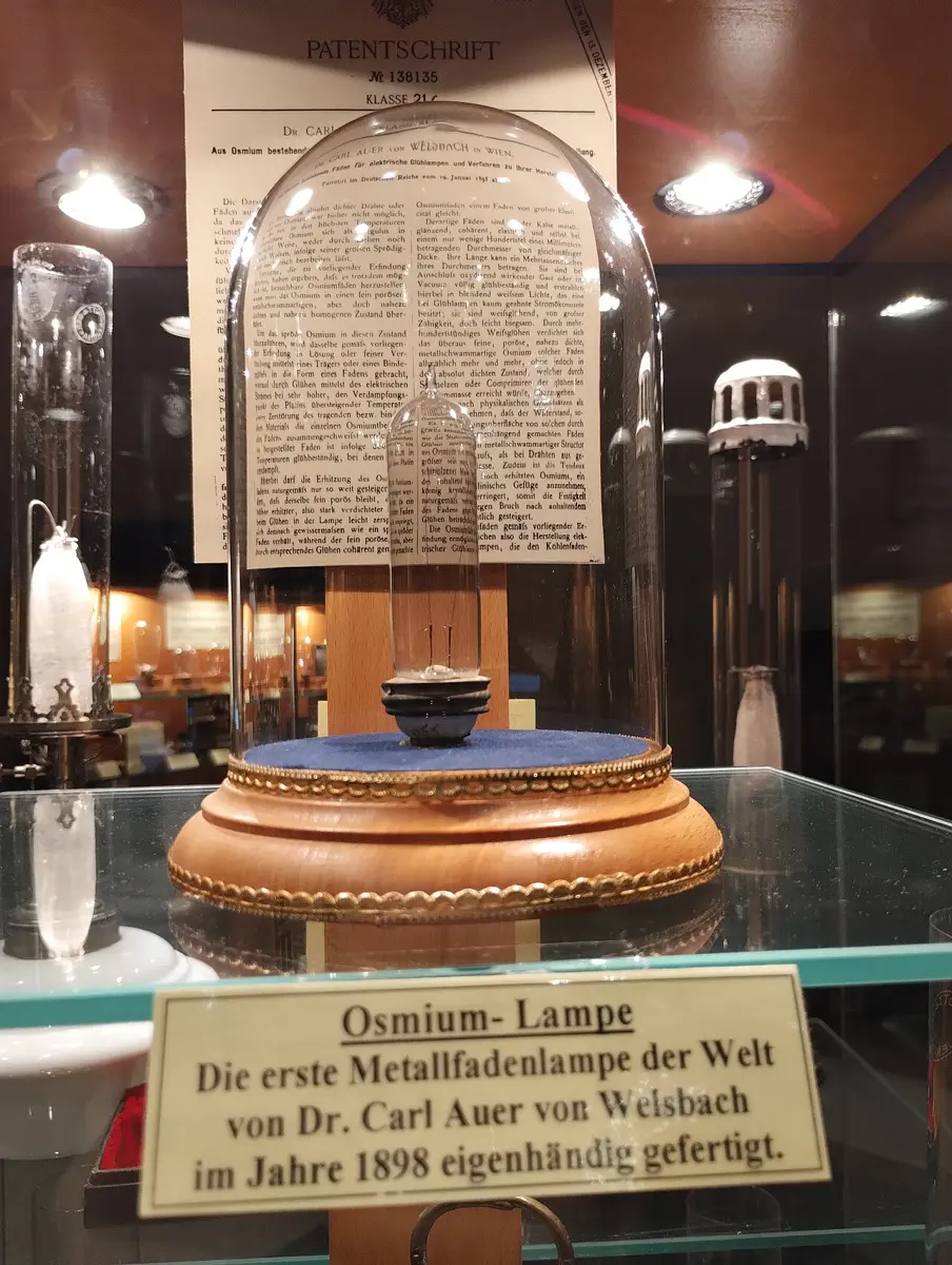 A small, old glass light bulb on a wooden stand under a glass dome in a museum case, with a printed patent behind it and a German label in front