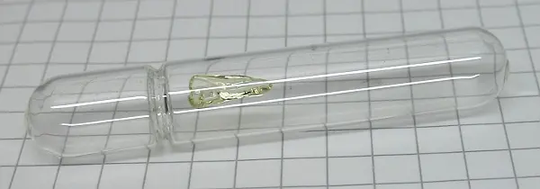 A sealed glass tube, like a small capsule, lying on graph paper, with a pale yellow-green crystal inside