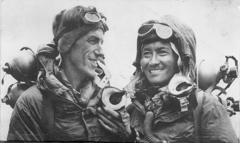 Two smiling climbers in hoods and goggles, with oxygen masks hanging under their chins and oxygen cylinders on their backs