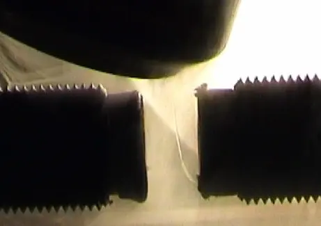 A thin stream of pale liquid falling past the gap between two dark, ridged magnet poles, bent sideways towards one of them