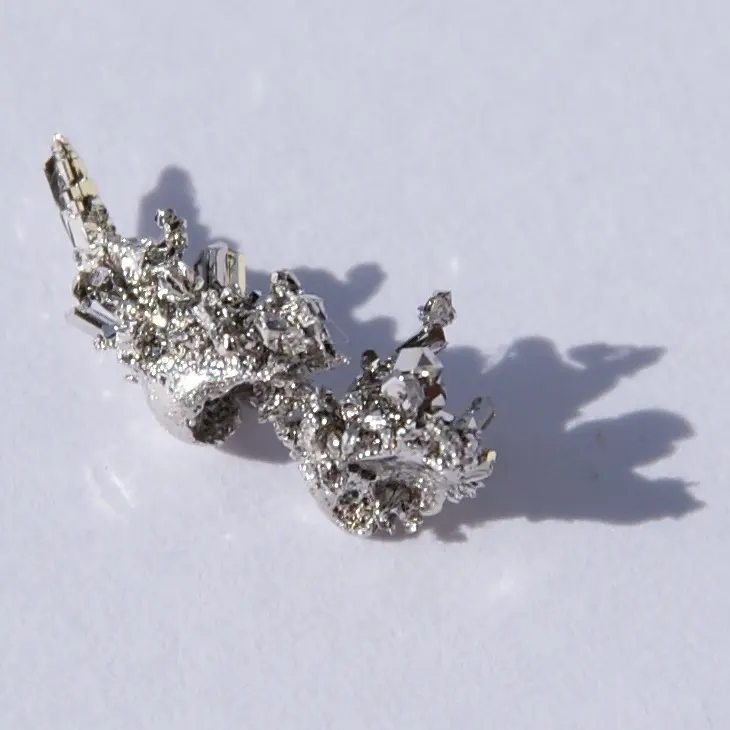A small, bright silvery piece of metal covered in tiny sharp crystals, casting a shadow on white paper