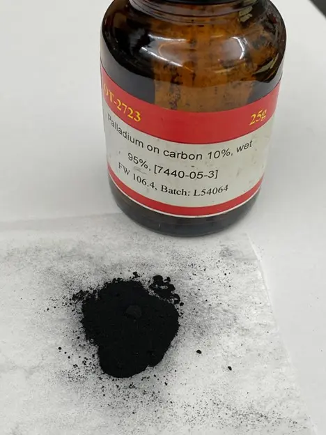 A small heap of black powder on white paper, in front of a brown glass bottle labeled Palladium on carbon 10%