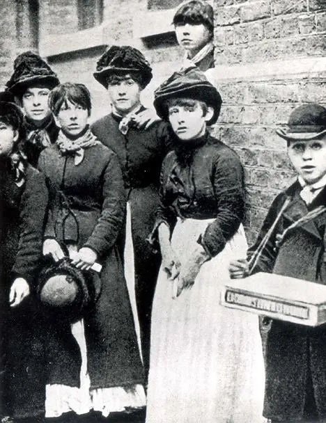 An old black-and-white photo of a group of young women and girls in long dark dresses, aprons and hats, standing against a brick wall; one holds a tray of match boxes