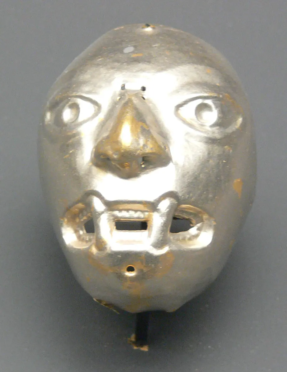 A small, pale silver mask of a face with round eyes, a large nose and an open mouth showing teeth, with a few gold-colored spots