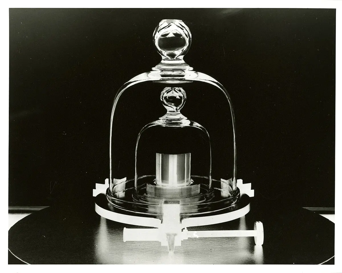 A black-and-white photo of a small, shiny metal cylinder standing under two glass bell jars, one inside the other, on a round base