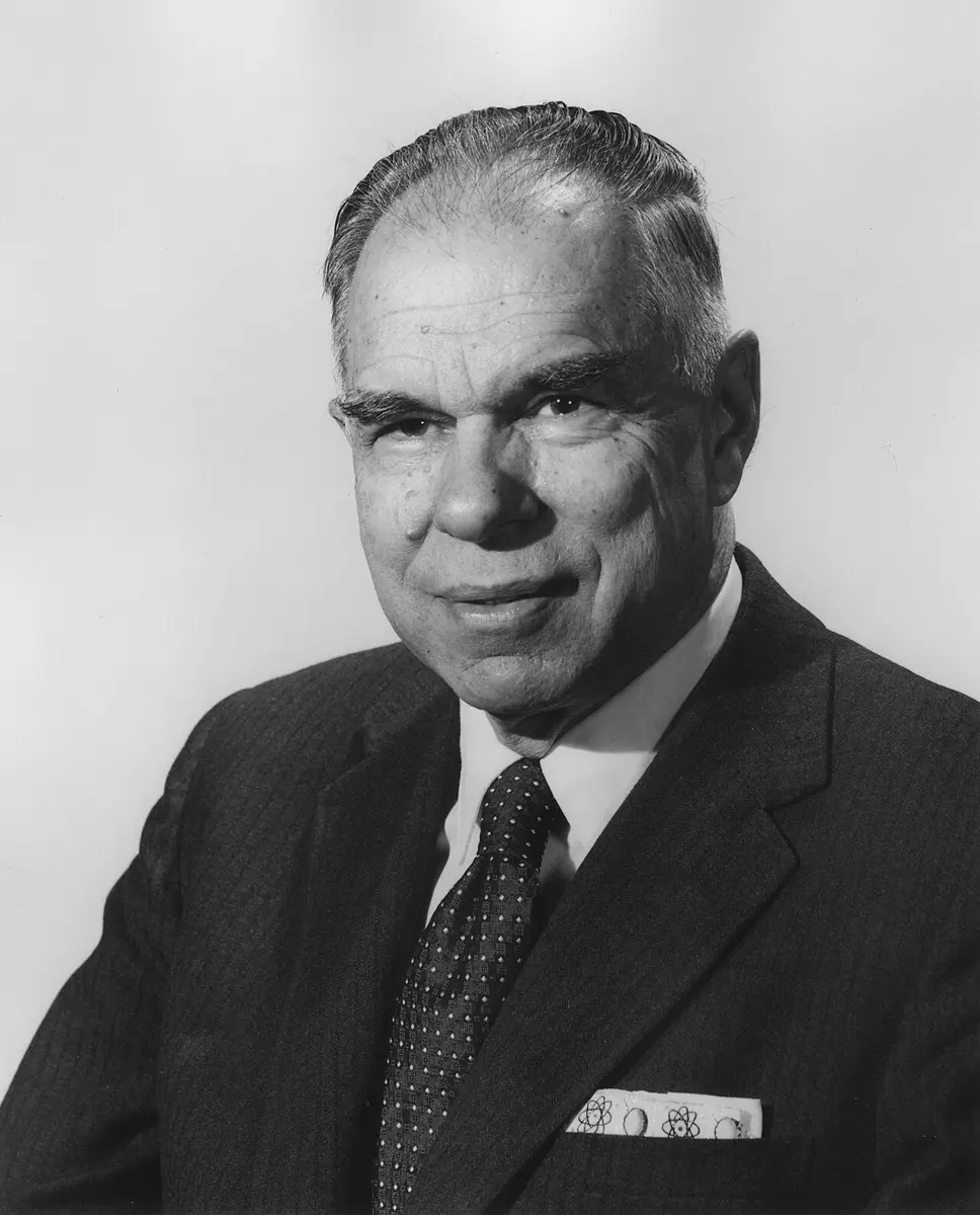 A black-and-white portrait of a smiling man with graying hair, in a dark suit, a patterned tie and a pocket handkerchief