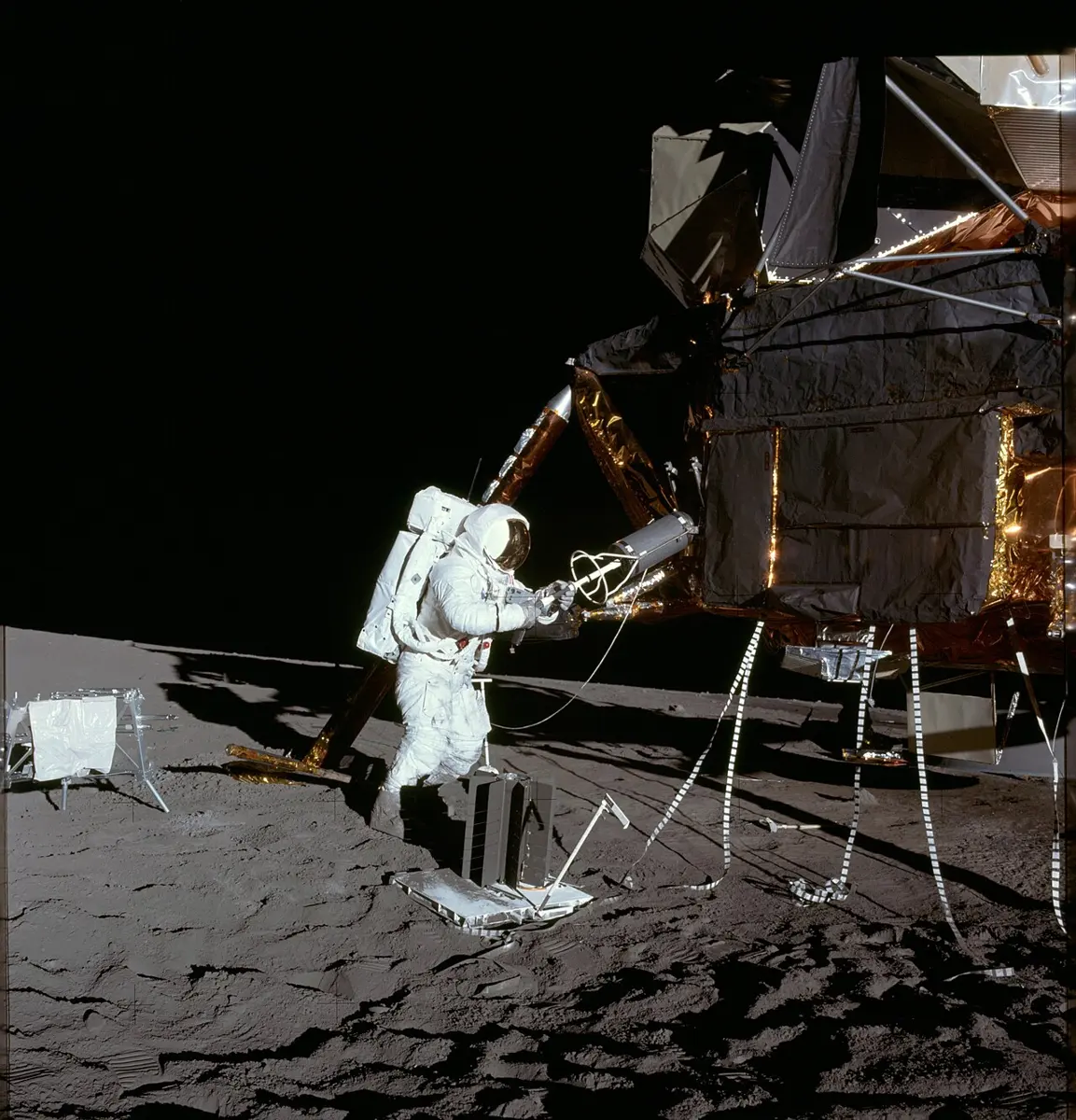 An astronaut in a white spacesuit on the gray surface of the Moon, beside the gold-foil-covered legs of a lunar lander, lifting a small cylinder out of a case on its side