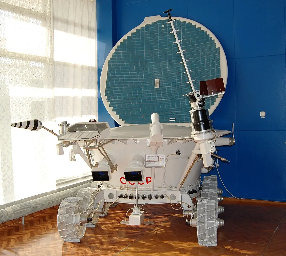 A white, tub-shaped vehicle on eight wire-mesh wheels, with cameras at the front and an antenna on a mast, in a museum room