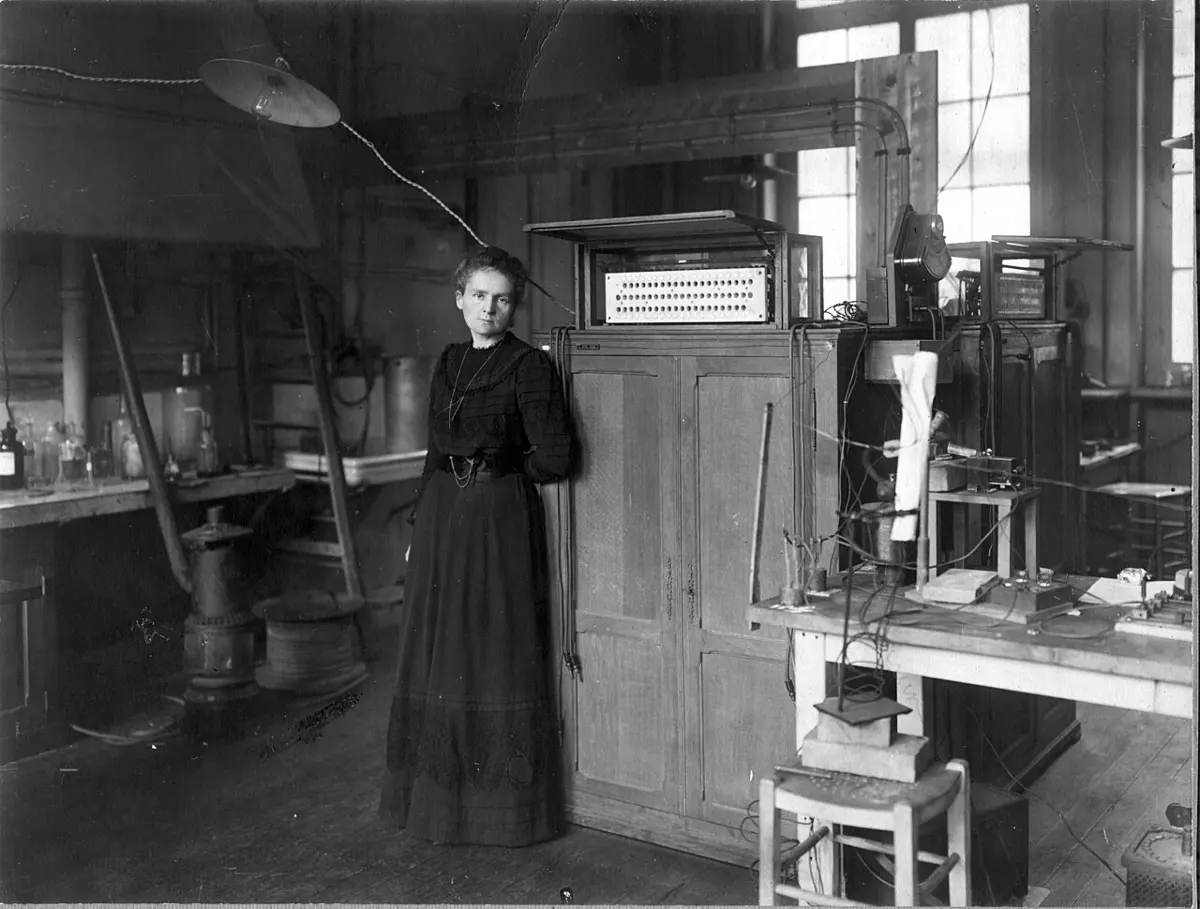 A black and white photograph of a woman in a long dark dress standing beside a tall wooden cabinet in a laboratory full of benches, glassware and instruments
