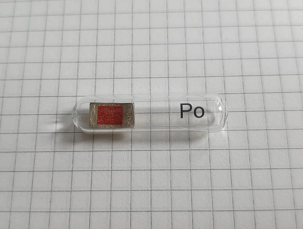 A small sealed glass tube on squared paper, marked Po, holding a tiny piece of brass with a reddish-brown patch