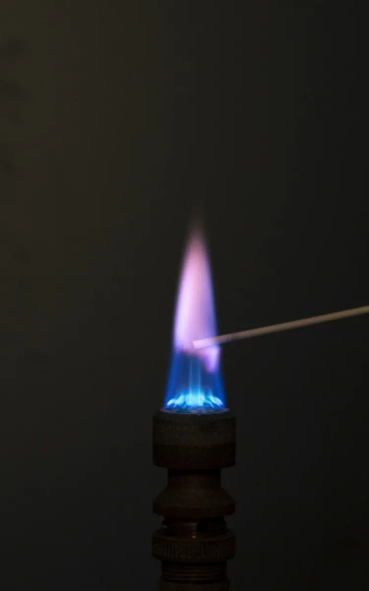 A blue gas flame in the dark with a thin wire held in it; above the wire the flame turns pale lilac