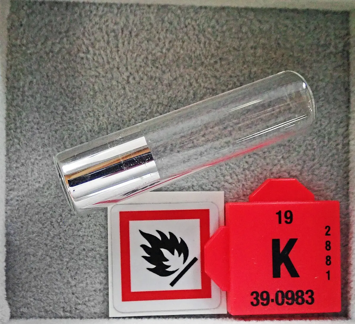 A sealed glass tube holding a shiny silver plug of metal, beside a red tile reading 19, K, 39.0983 and a flammable-hazard sign