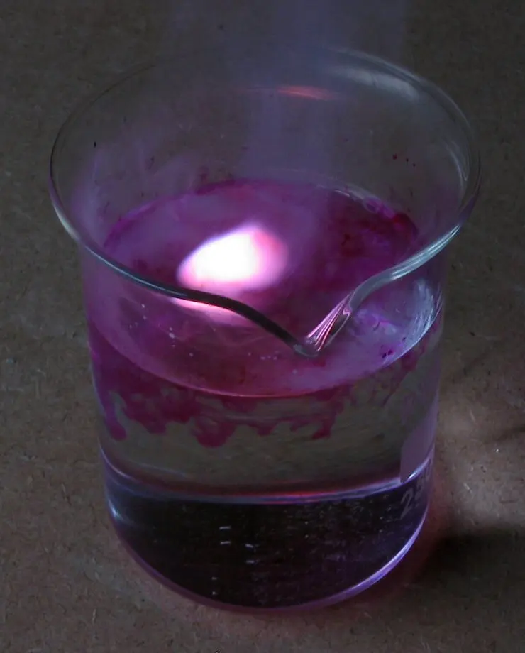 A glass beaker of water with a small lilac flame on its surface and pink streaks spreading down through the water