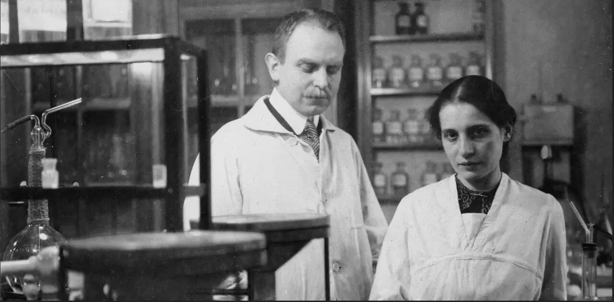 An old black-and-white photograph of a man with a mustache and a young woman with dark hair, both in white laboratory coats, standing among glassware and shelves of bottles
