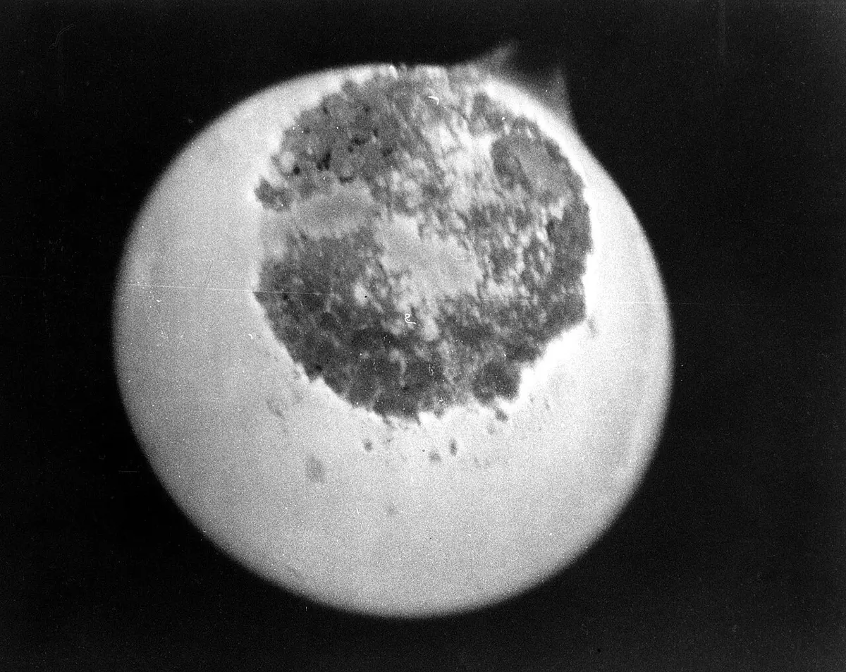 A black-and-white photo of a glowing pale disc on a black background, with a dark, grainy patch covering its upper middle