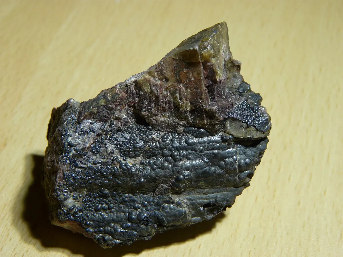 A lump of rock on a wooden table, its lower part covered in shiny, dark gray-black bubbles, with a brownish crystal on top