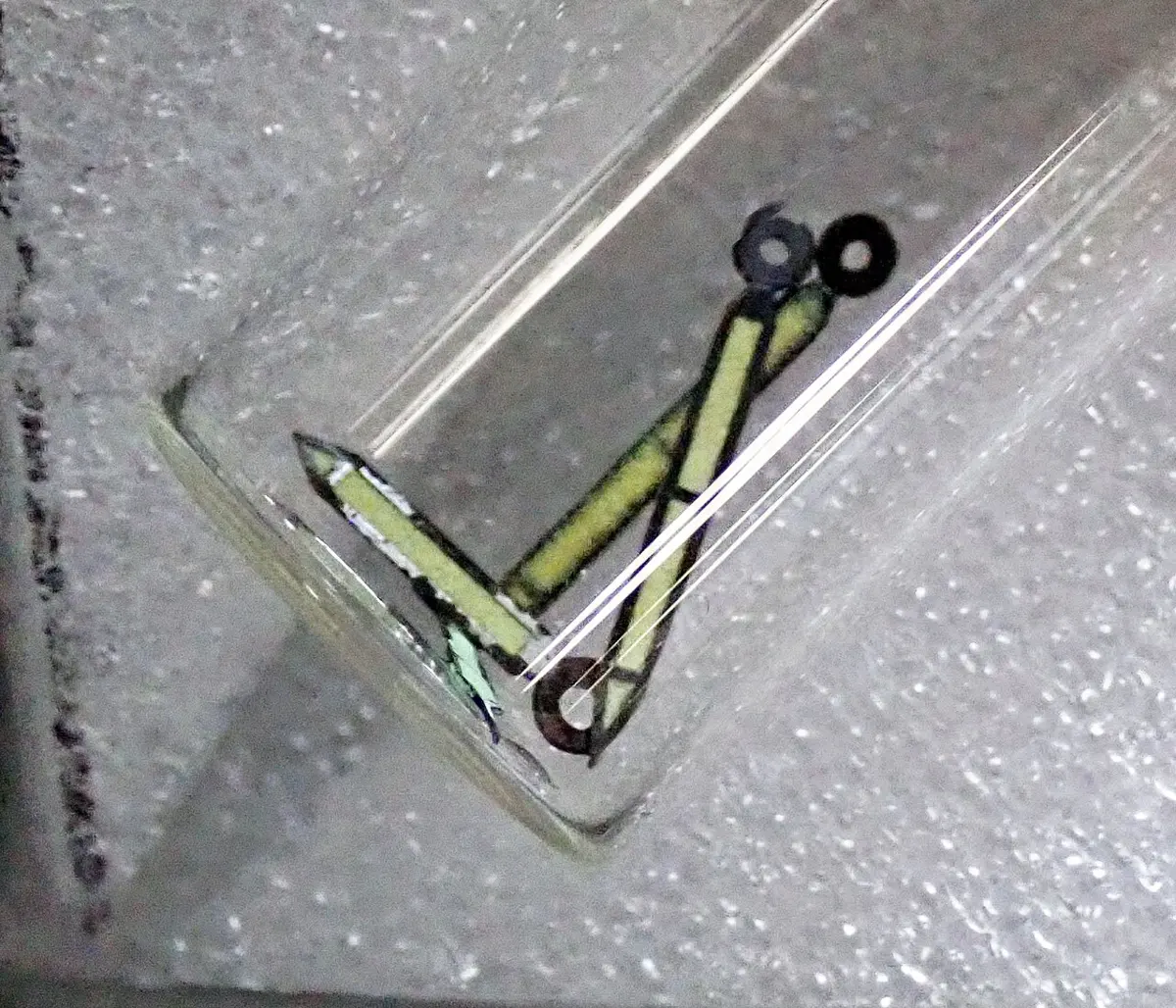 Three thin watch hands with black edges and pale yellow-green paint inside them, in a small glass tube