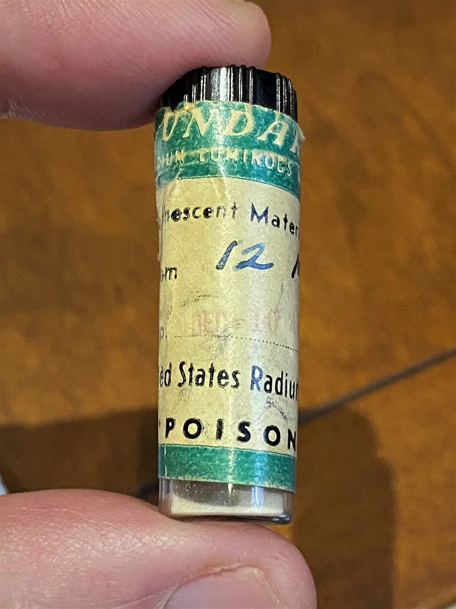 Fingers holding a small glass tube with a black cap and an old paper label reading Undark, United States Radium, Poison, with a pale powder at the bottom