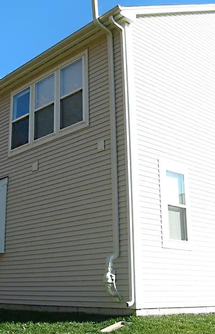 The corner of a two-storey house with pale siding; a white plastic pipe runs from a small box near the ground all the way up the wall to above the roof