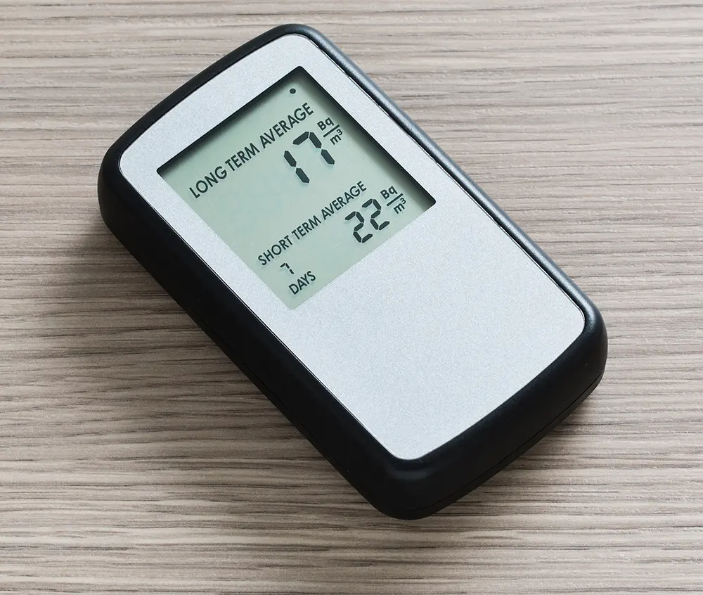 A small gray electronic device with a black rim on a wooden table, its screen showing LONG TERM AVERAGE 17 Bq/m3 and SHORT TERM AVERAGE 22 Bq/m3, 7 days