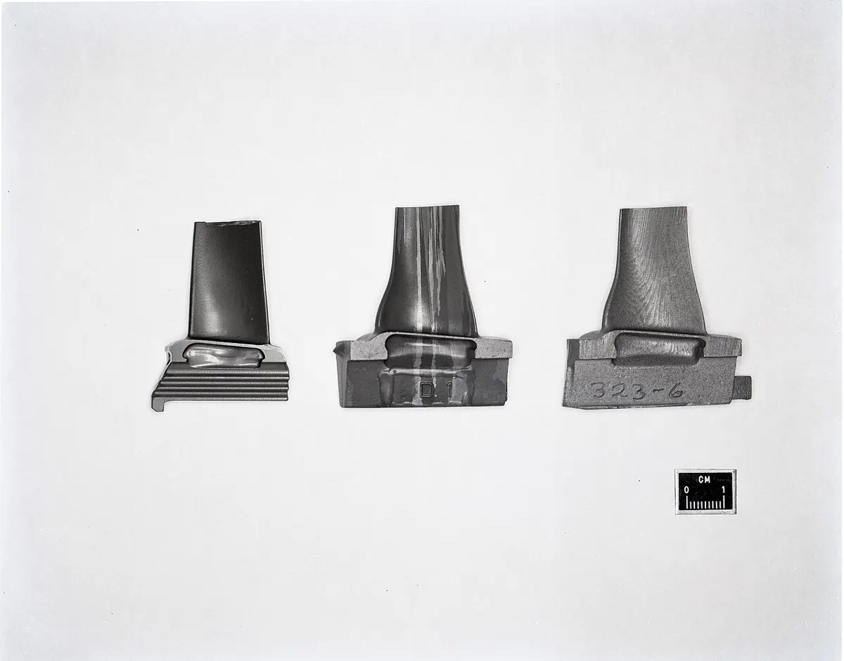 Three dark gray metal turbine blades standing on their roots, each a few centimeters tall, with a small centimeter scale beside them