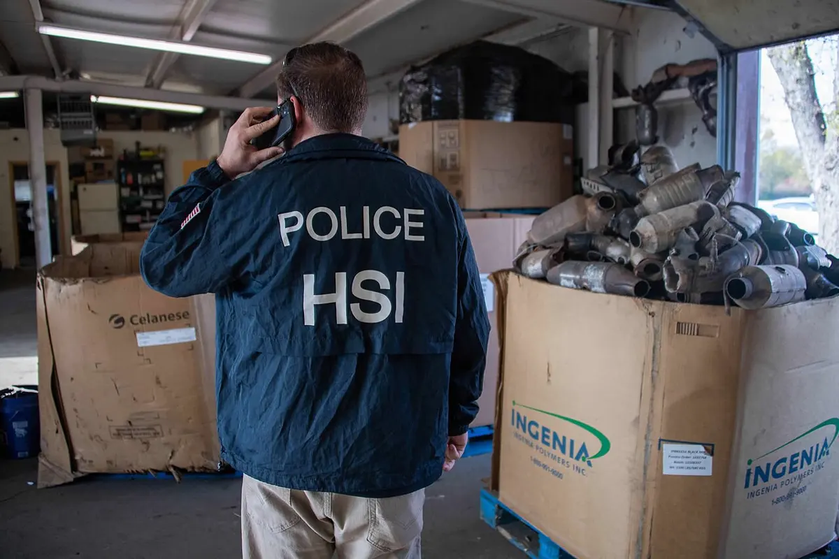 A police officer, seen from behind in a jacket marked POLICE HSI, beside a large cardboard box piled high with metal exhaust parts