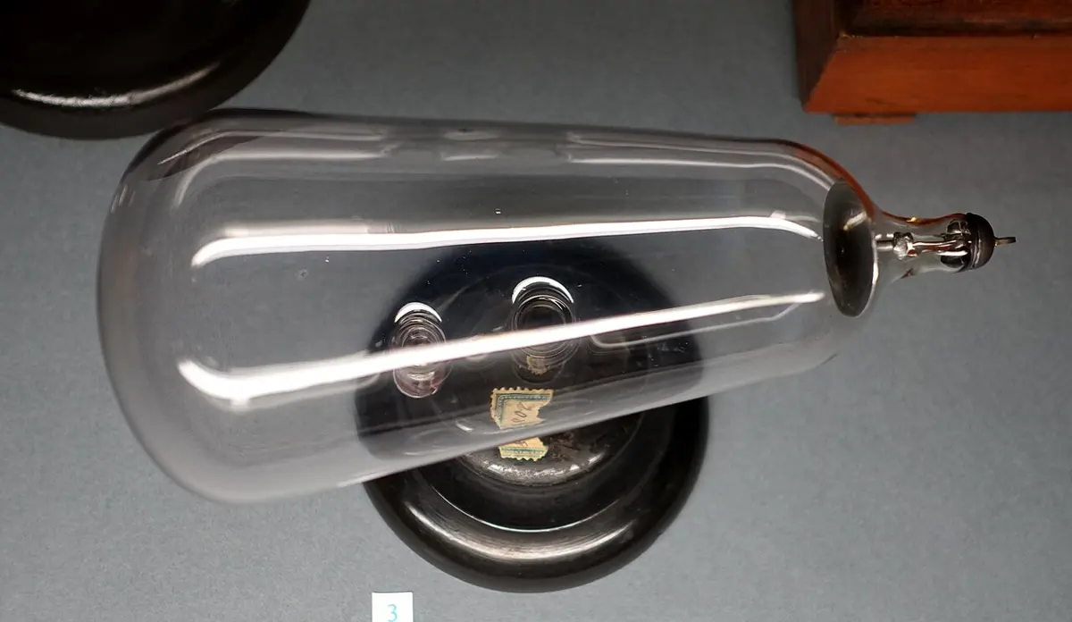 A pear-shaped clear glass tube lying on its side on a dark round stand, with a metal contact sealed into its narrow end and a numbered museum label in front