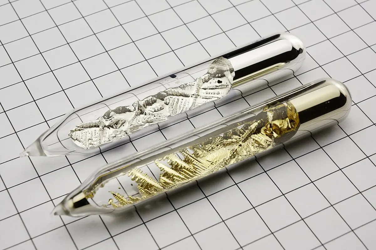 Two sealed glass tubes on squared paper: the upper one holds bright silvery crystals, the lower one golden crystals