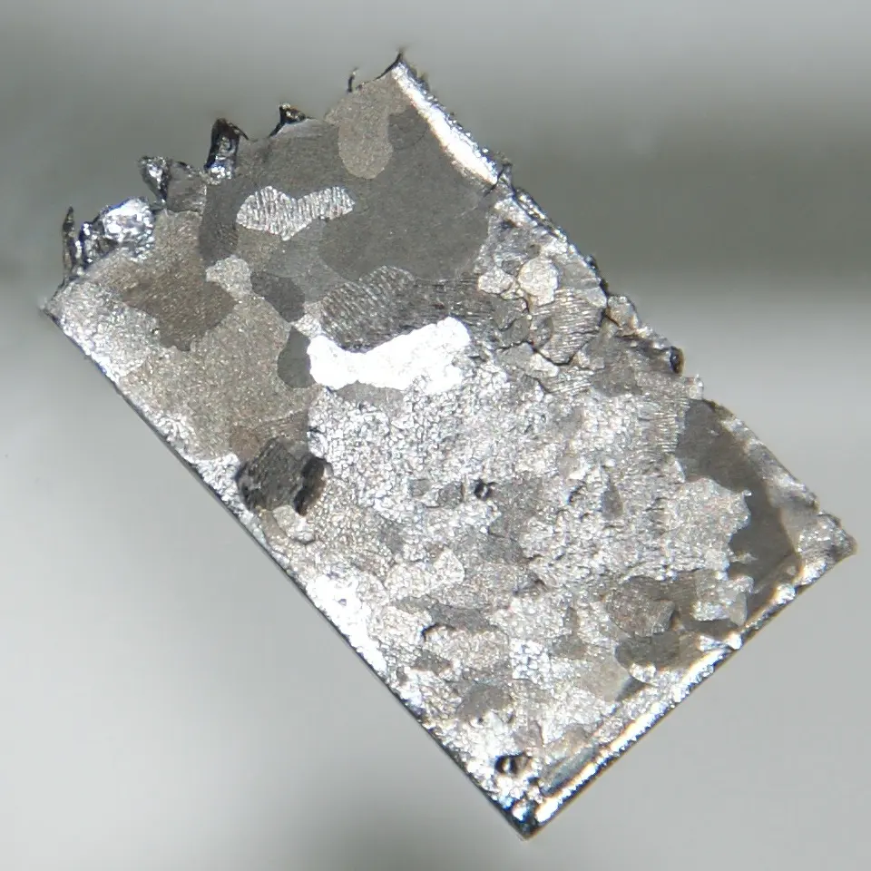 A small, flat, rectangular piece of silvery metal with a jagged edge at one end; its surface shows a patchwork of lighter and darker grains