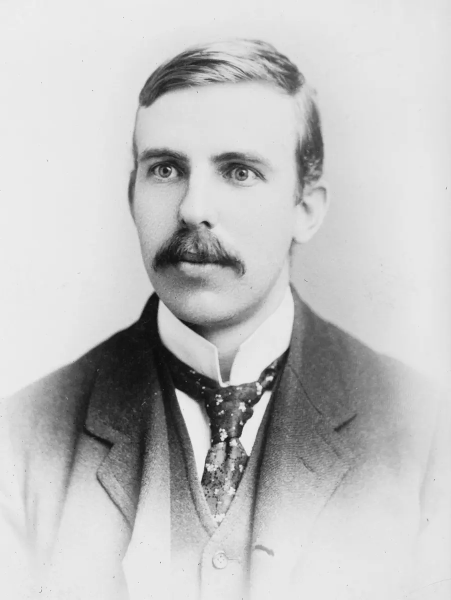 A black-and-white portrait of a young man with short hair and a thick mustache, in a high collar, patterned tie and waistcoat