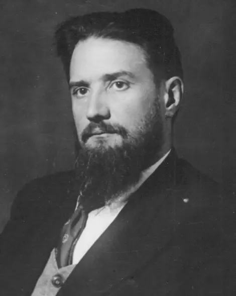 A black-and-white portrait of a man with swept-back dark hair and a long, full beard, in a jacket and tie