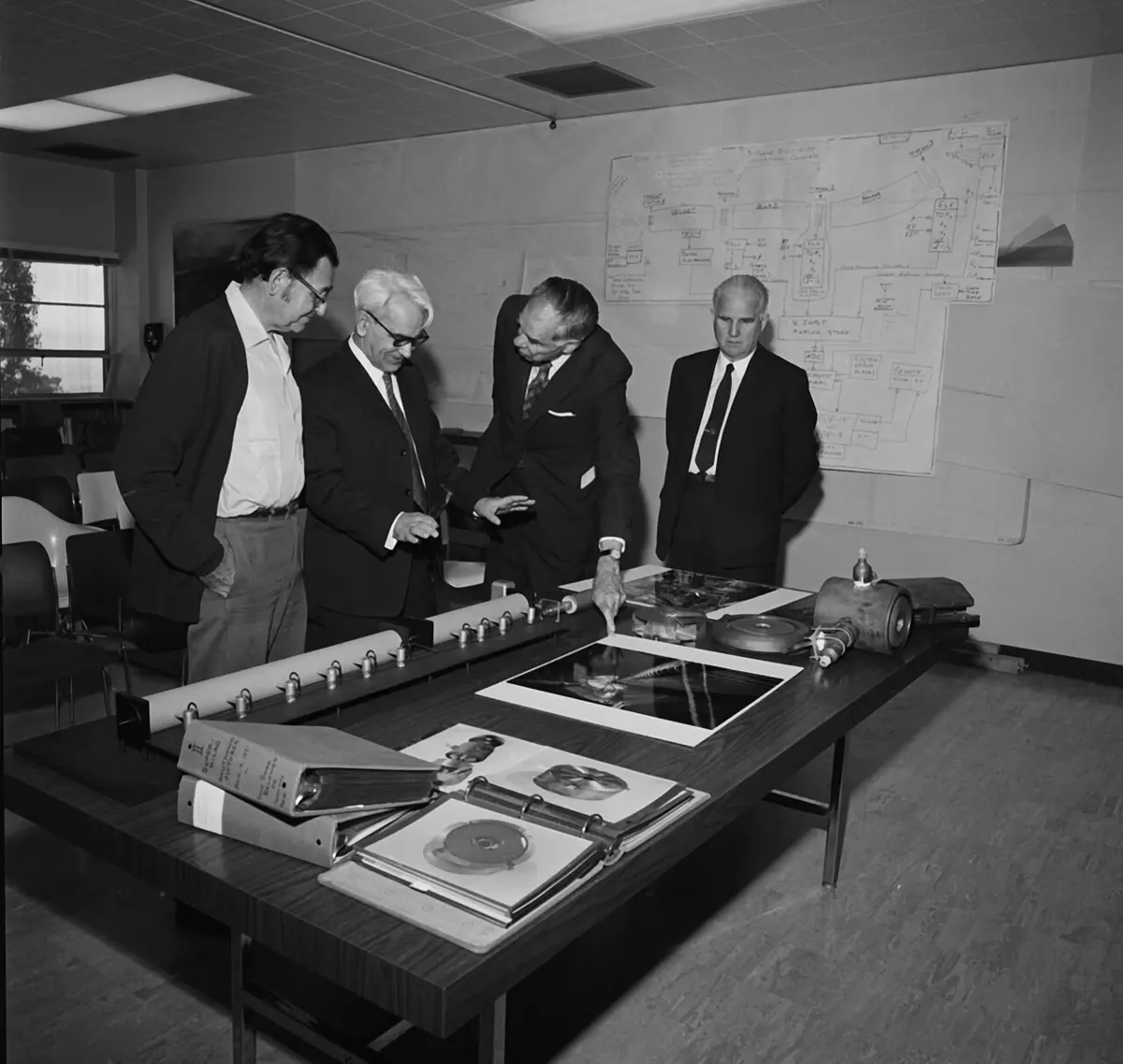 A black-and-white photo of four men in suits standing at a table covered with photographs, folders and rolled-up charts, one pointing at a photograph, with a wiring diagram pinned on the wall behind